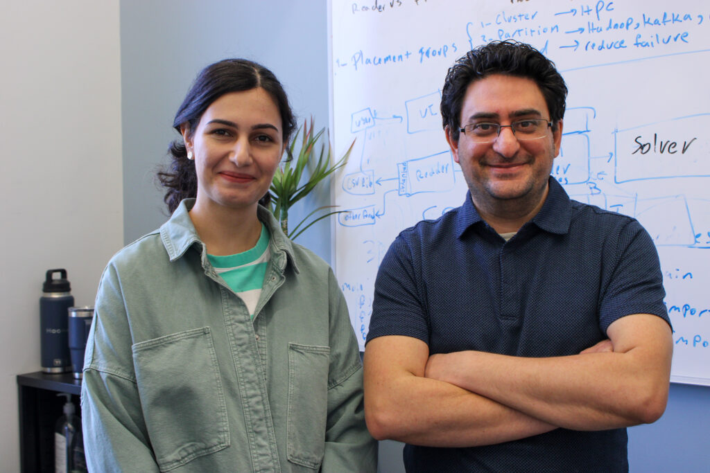 Sogand Soghrati and Hooman Niktafar smiling at the camera while standing in front of a whiteboard with writing on it.