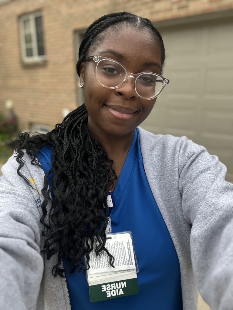 Chidimma wearing a blue scrub top, gray zip sweatshirt, and backwards "Nurse Aide" badge.