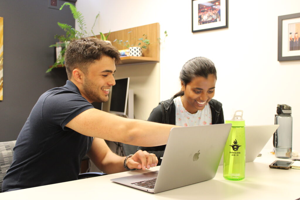 Arman smiling and pointing to an open laptop beside another student.