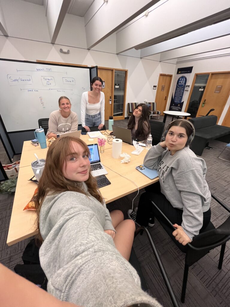 Hannah and four other CHEPS students smiling for a selfie while woring at a large table.