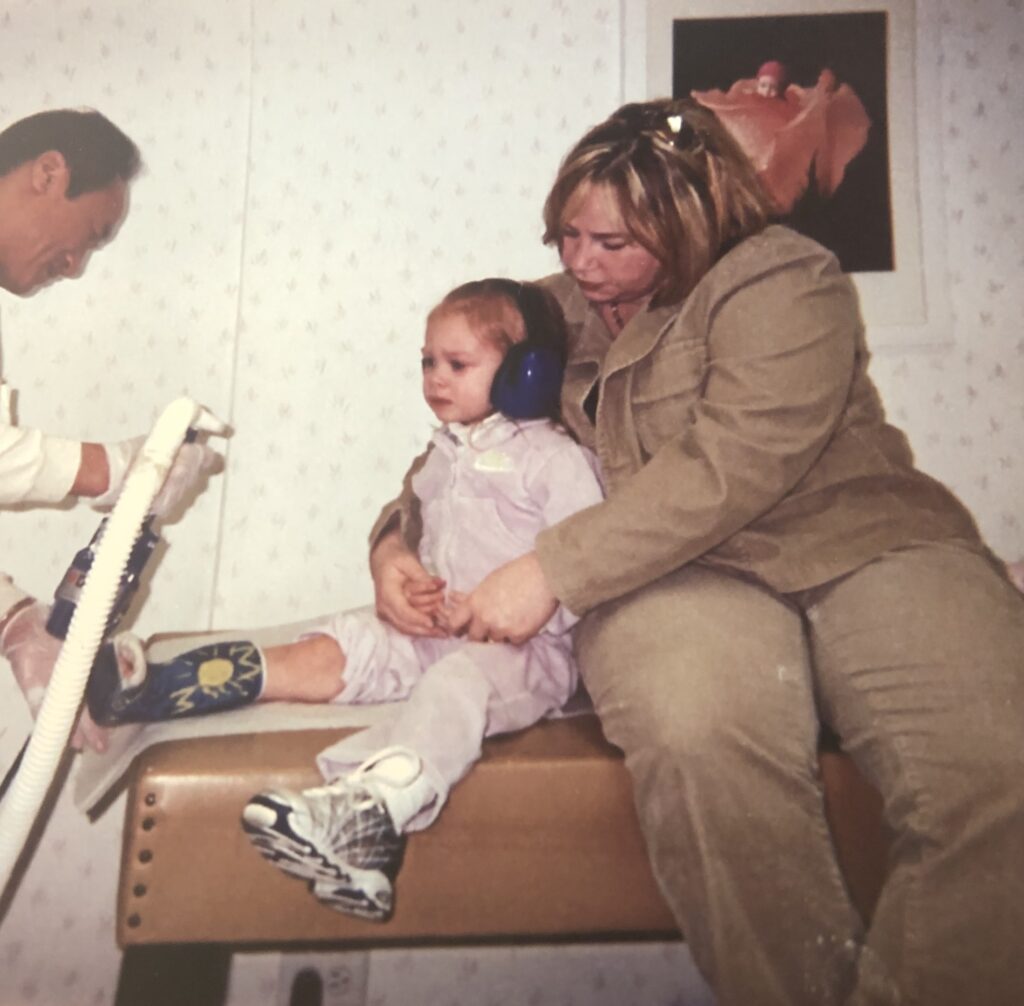 A young Hannah getting a cast removed while wearing over-ear headphones and being held by her mother.