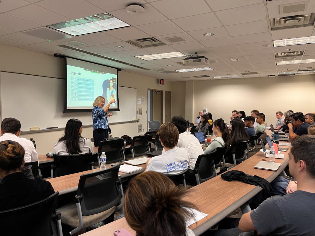 Amy Cohn wewaring a blue blouse and presenting in front of a small lecture hall, gesturing out towards the audience while standing in front of a projected screen reading "Agenda."