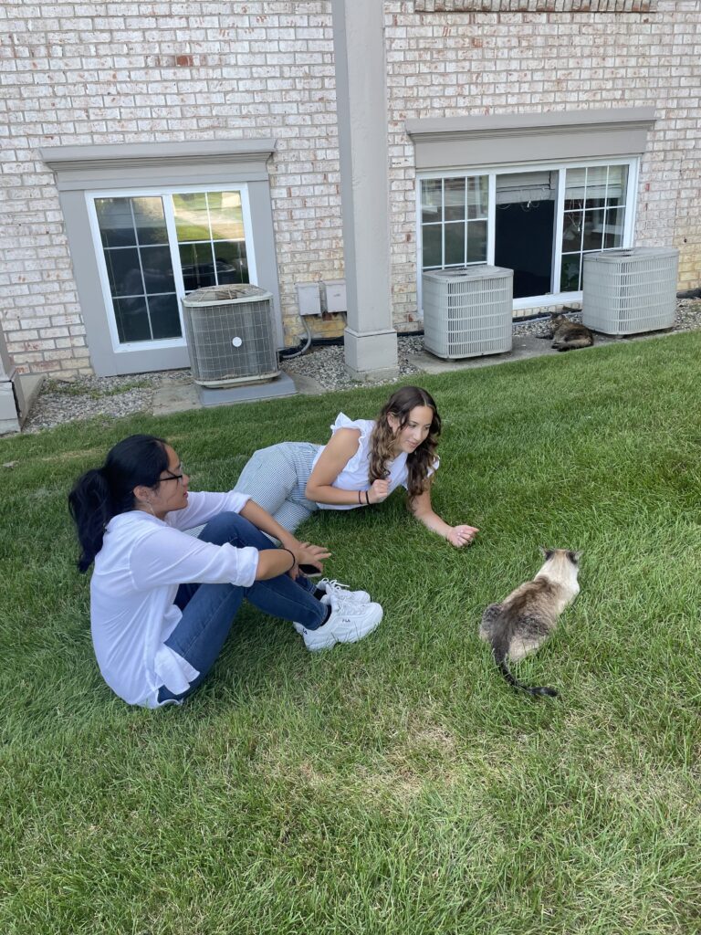 Jill and Goretti sitting in the grass and giving attention to a brown cat.