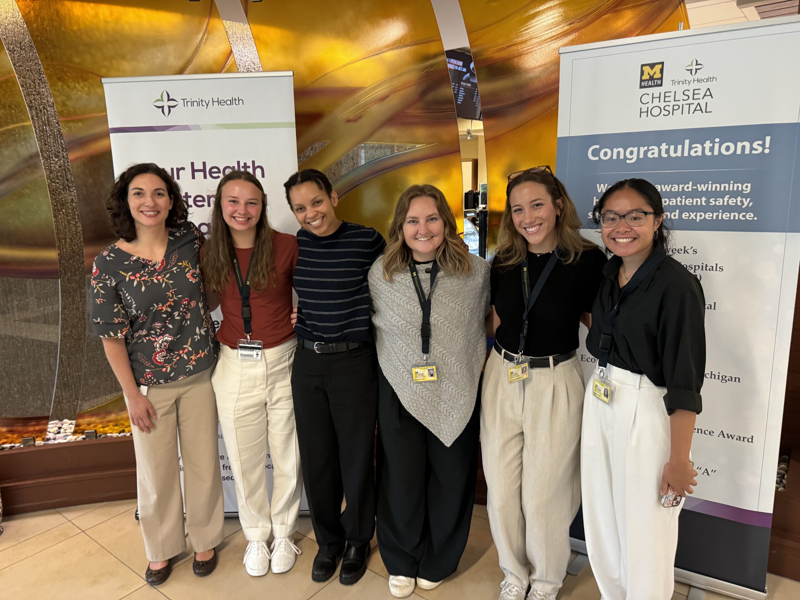 Six women (members of the Clinical Research Ethnography team) stand in a line and smile. They wear professional dress and are in front of hospital signage.